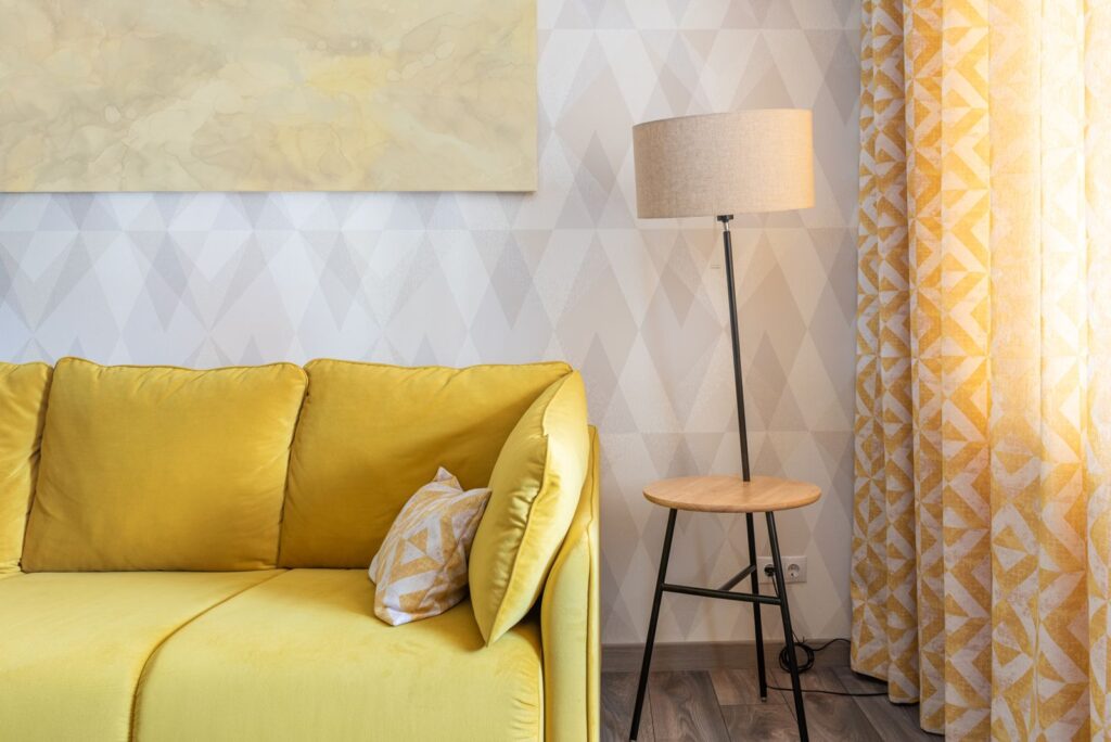 Rental living room with patterned accent wallpaper, a floor lamp and floor-length curtains behind a yellow sofa